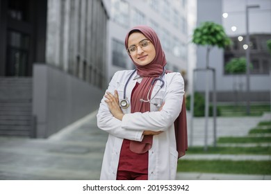Arab Female Doctor Or Medical Student, Posing And Smiling With Folded Arms, Standing Outside On A Background Of Modern University Campus Or Hospital Building