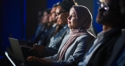 Arab Female Sitting In A Dark Crowded Auditorium At A Human Rights Conference. Young Muslim Woman Using Laptop Computer. Activist In Hijab Listening To Inspiring Speech About Global Initiative.