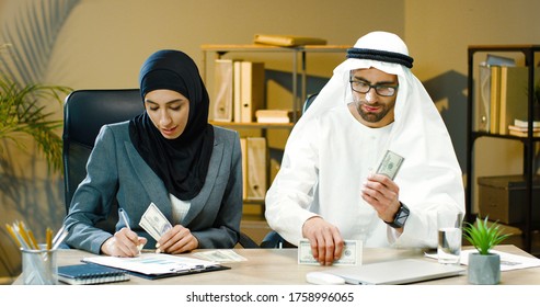 Arab Girl On Abaya Hijab Black Dress And Arab Man Counting Money American Dollars. Arab Saudi Man And Beautiful Arabic Emirati Lady With Cash. 