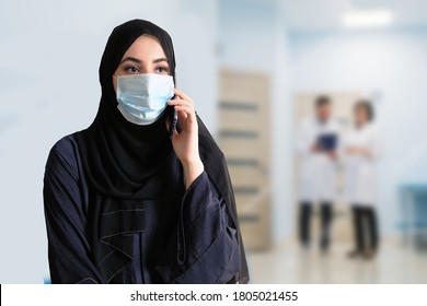 Arab At Hospital Lobby Wearing Protective Face Mask With Blurred Doctors At The Background. Emirati Woman Using Mobile Phone As She Calls