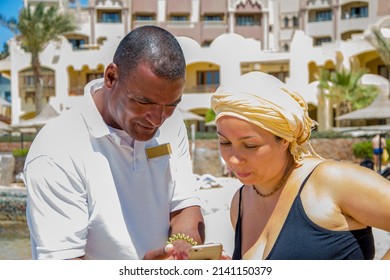 An Arab Hotel Employee Shows Something On The Phone To A 40-year-old Woman In A Swimsuit On The Beach.