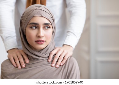 Arab Husband Threatening Scared Wife In Hijab Touching Her Shoulders Standing Behind Her Back At Home. Domestic Violence And Abuse, Unhappy Marriage With Abuser Problem. Cropped, Selective Focus