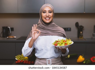 Arab Lady Cooking Standing In Modern Kitchen, Holding Salad Bowl Gesturing OK And Smiling To Camera, Wearing Hijab At Home. Dinner Preparation, Female Muslim Restaurant Chef Approving Food And Recipe