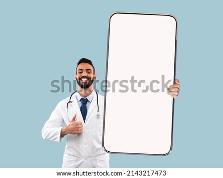 Arab male doctor showing smartphone with white blank screen and gesturing thumb up over blue studio background. Medical worker demonstrating mobile phone screen, mockup
