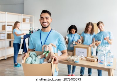 Arab male volunteer holding box with clothes and toys for donation, smiling to camera while working at charity center with group of colleagues, packing products in boxes on background - Powered by Shutterstock