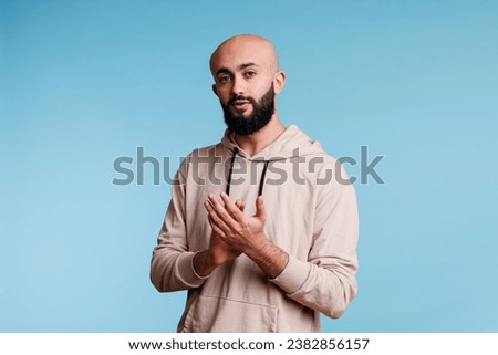 Arab man applauding with hands while standing and looking at camera with neutral expression. Young bald bearded person clapping with arms, congratulating and supporting studio portrait