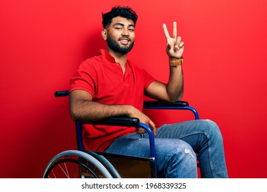 Arab man with beard sitting on wheelchair smiling with happy face winking at the camera doing victory sign with fingers. number two. 