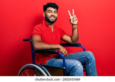 Arab man with beard sitting on wheelchair smiling looking to the camera showing fingers doing victory sign. number two. 