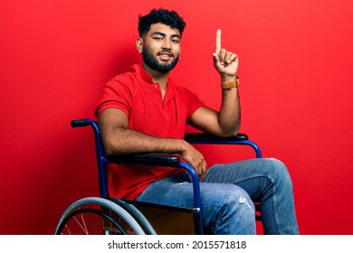 Arab man with beard sitting on wheelchair pointing finger up with successful idea. exited and happy. number one. 