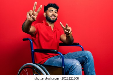 Arab man with beard sitting on wheelchair smiling with tongue out showing fingers of both hands doing victory sign. number two. 