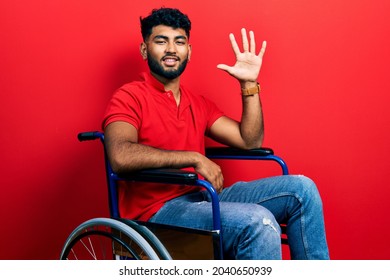 Arab man with beard sitting on wheelchair showing and pointing up with fingers number five while smiling confident and happy. 