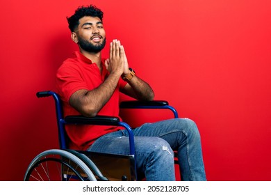 Arab Man With Beard Sitting On Wheelchair Praying With Hands Together Asking For Forgiveness Smiling Confident. 