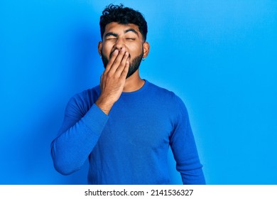 Arab man with beard wearing casual blue sweater bored yawning tired covering mouth with hand. restless and sleepiness. 