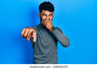 Arab Man With Beard Wearing Turtleneck Sweater Laughing At You, Pointing Finger To The Camera With Hand Over Mouth, Shame Expression 