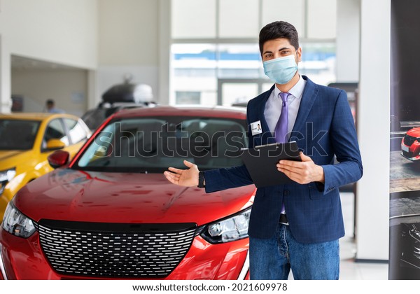 Arab man in formal outfit and protective face mask\
standing over luxury red car in big showroom, copy space. Male\
sales manager pointing at brand new automobile, holding chart with\
info about auto