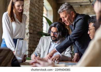 Arab man signs a contract guided by an older man in a suit while a diverse group of people look on. Multicultural team works together to finalize a business deal, diversity and collaboration concept - Powered by Shutterstock