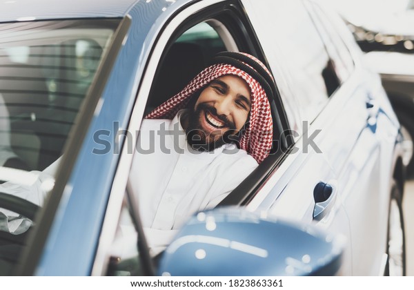 Arab man sits at the wheel of a car and looks\
out of the window. Arab man is testing a car at a car dealership\
and smiling.