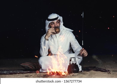 Arab Man Sitting Close To Bonfire In The Desert At Night Drinking Tea Wearing Traditional Clothes 