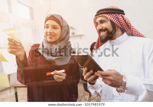 タウブを着たアラブ人の男性とヒジャブの女性がオフィスで働いている 同僚がガラス板にメモを書いている の写真素材 今すぐ編集