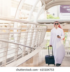 Arab Man In Travel Concept . Young Saudi Arabic Man In Traditional Clothes Walking With Suitcase On Airport Background