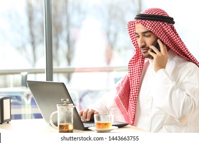 Arab Man Using A Laptop And Talking On Snmart Phone Sitting In A Coffee Shop