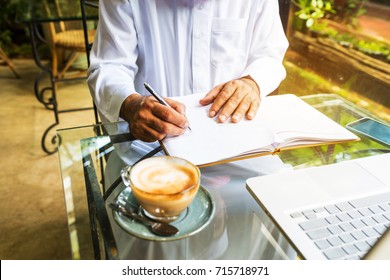 Arab Muslim Business Man Ware White Traditional Clothing And Write On Paper ,On Table Have Laptop Smartphone And Cup Of Coffee