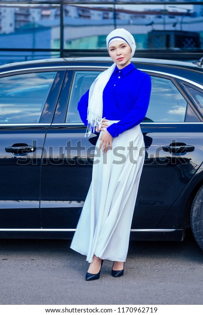 Arab Muslim business woman\
hijab with makeup standing in front of her luxury car on the street\
on a background of skyscrapers . The woman is dressed in a stylish\
abaya