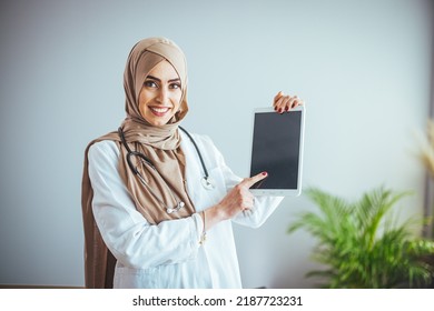 An Arab Muslim Trainee Doctor Smiles At An Isolated White Background Concept For Islamic People Working In A Medical Hospital, Pointing To A Digital Tablet. The Concept Of Modern Technology And Health