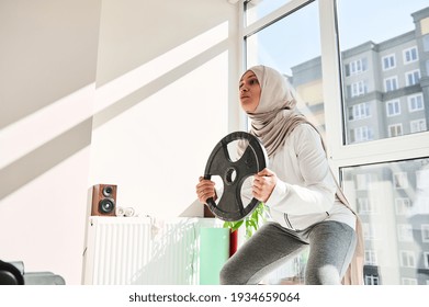 Arab Muslim Woman With Headdress Wearing Hijab Doing Weighted Squats With A Metal Disc In Her Hands - Home Workout Concept.
