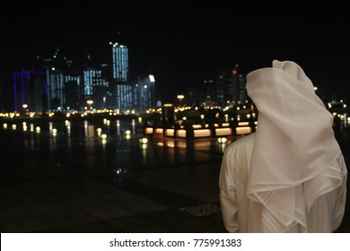 An Arab Qatari Man Wearing A White Thobe Or Thwab (Arab Garment) And White Ghutra (or Keffiyeh Or Headress) Looking Out To The Evening Or Night Skyline Of Doha, Qatar