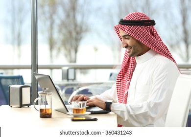 Arab Saudi Man Working Online With A Laptop And Smartwatch In A Coffee Shop Or An Hotel Bar With A Window And Outdoor Terrace In The Background