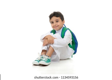 Arab school boy sitting on ground with a smile on his face, wearing white traditional Saudi Thobe, back pack and sneakers, raising his hands on white isolated background