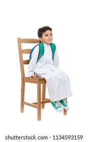 Arab school boy sitting on a wooden chair with a smile on his face, wearing white traditional Saudi Thobe, back pack and sneakers, raising his hands on white isolated background