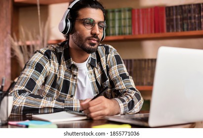 Arab Student Guy Learning Online At Laptop Computer Studying Sitting In Library Near Bookshelf In University. Modern Education, E-Learning And Online Training Concept. Selective Focus