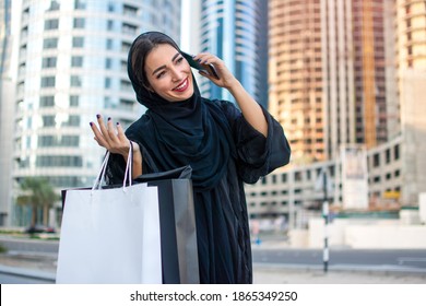 Arab Woman In Abaya Talking On Mobile Phone While Holding Shopping Bags And Walking On City Street.