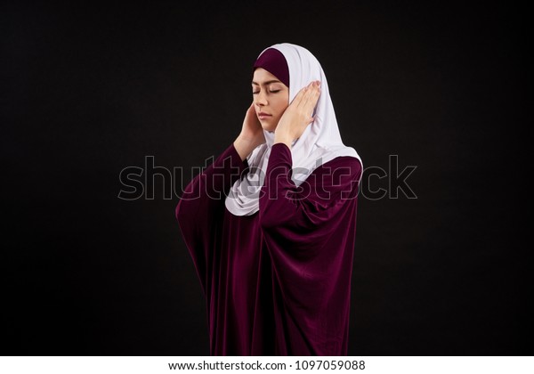Arab woman in hijab covers ears with\
hands. Hear no evil. Isolated on black\
background.