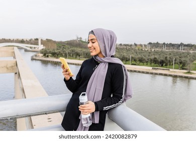 Arab woman rests from exercise looking at her cell phone and refreshing herself with water - Powered by Shutterstock