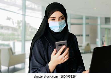 Arab Woman Wearing Face Mask While Using Mobile Smart Phone And Practicing Social Distance At Office. Emirati Arabic Lady On Traditional Hijab Abaya As Part Of Middle East Culture