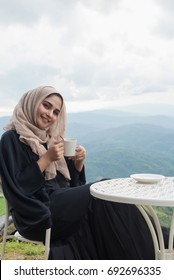 Arab Women In Hijab Holding Coffee Sitting On The Background Of Mountain And Looking At Camera. Woman Is Dressed In A Black And  Scarf Brown.