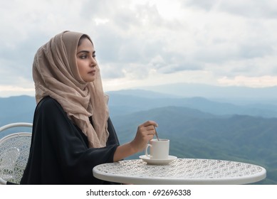 Arab Women In Hijab Holding Coffee Sitting On The Background Of Mountain And Looking At Camera. Woman Is Dressed In A Black And  Scarf Brown.