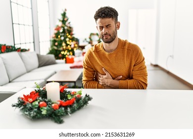 Arab Young Man Sitting On The Table By Christmas Tree With Hand On Stomach Because Indigestion, Painful Illness Feeling Unwell. Ache Concept. 