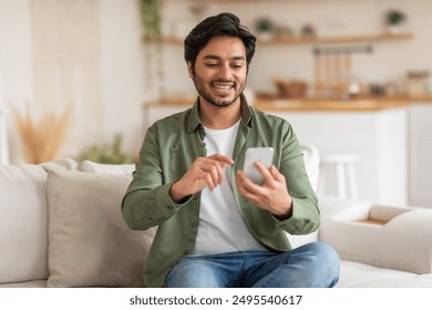 Arab young man, wearing a green shirt and jeans, smiles while looking at his phone. He is sitting on a white couch in a modern home. - Powered by Shutterstock