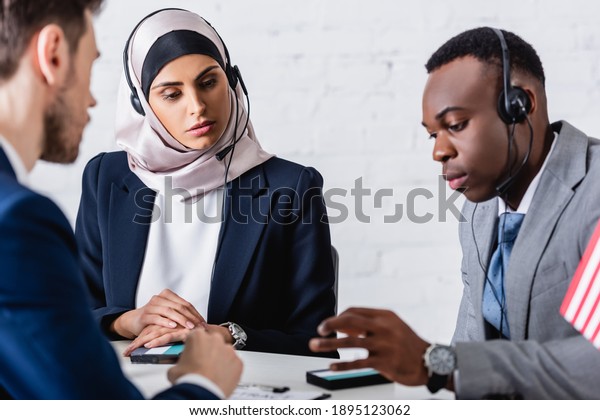 arabian\
and african american business people in headsets near digital\
translators and interpreter on blurred\
foreground