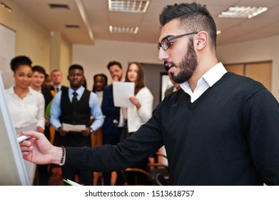 Arabian business coach presenting report standing near whiteboard pointing on sales statistic shown on diagram and chart teach diverse multiracial company members gathered together in conference room.