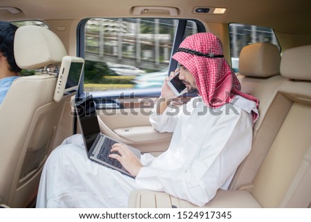 Arabian businessman working with laptop while making a phone call and sitting backseat in a car 