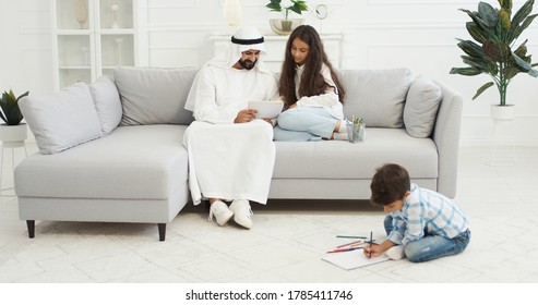 Arabian Father In White Kandura Playing With Small Kids, Daughter And Son In Living Room. Muslim Dad Drawing And Coloring Pictures. Boy And Girl With Daddy Painting At Home And Spending Time Together.