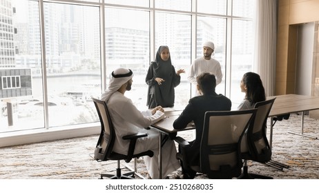 Arabian female manager in hijab and abaya presenting business project strategy to team of diverse colleagues, standing at speaking to Arab and European colleagues at large meeting table - Powered by Shutterstock