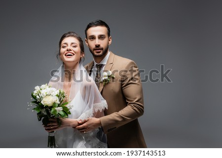 Arabian groom embracing cheerful bride with bouquet isolated on grey