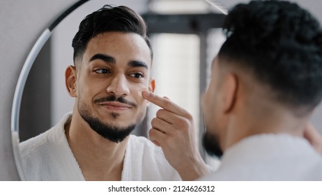 Arabian Indian Arab Hispanic Millennial 30s Bearded Man Applying Moisturizer Cream On Face Skin Looking In Bathroom Mirror In Morning Guy Puts Facial Creme Doing Skincare Beauty Hydration Routine