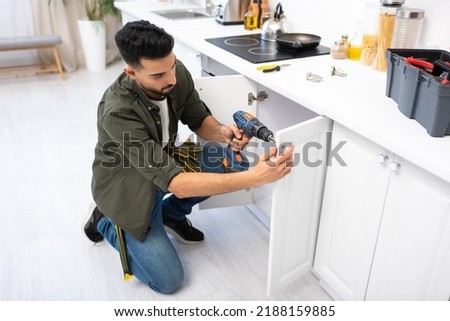Arabian man with electric screwdriver fixing cabinet under worktop in kitchen